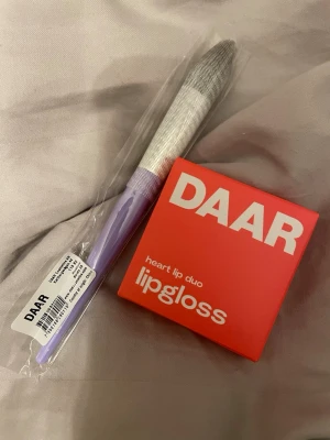  pocket beating lipgloss duo DAAR vanilla & strawberry + borste - Säljer ett DAAR heart lip duo lipgloss i smakerna vanilla & strawberry. Kommer i en snygg röd förpackning och passar perfekt för dig som vill ha glansiga och väldoftande läppar. Medf ne lila och vit applikatorborste i plast och syntet.