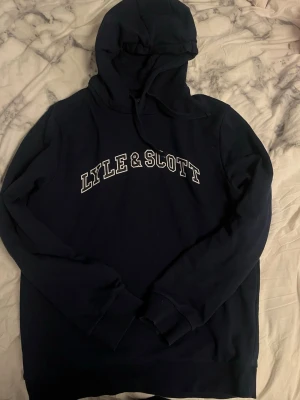 Marinblå hoodie från Lyle & Scott - Snygg marinblå hoodie från Lyle & Scott med vit logga tryckt över bröstet. Klassisk modell med huva och dragsko samt ribbade muddar vid ärmslut och nederkant. Perfekt för en avslappnad och stilren look. Tillverkad i mjukt bomullsmaterial.