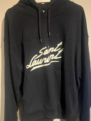 Svart hoodie från Saint Laurent - Snygg svart hoodie från Saint Laurent med vit logga i graffiti-stil på bröstet. Tillverkad i mjukt bomullsmaterial som känns riktigt skönt mot huden. Perfekt för dig som gillar streetwear och exklusiva märken. Pris är diskuterbart, hör av er om ni har frågor!