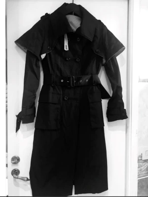 Svart trenchcoat/kappa med bälte och fickor - Stilren svart trenchcoat med dubbelknäppning, brett slag och bälte i midjan. Jackan har stora fickor framtill och knytdetaljer vid ärmsluten. Insidan är fodrad med ett ljust tyg med små prickar. Perfekt för dig som gillar klassisk men edgy stil.avtagbara axel partier 