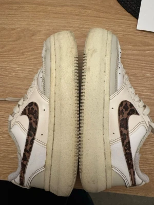Nike vita sneakers med leopardmönster - Säljer ett par vita Nike sneakers med klassisk låg siluett och leopardmönstrad Swoosh på sidan. Skorna har vita snören, perforerad tåbox och beige sula. Tillverkade i läder med detaljer i textil. Perfekta för dig som vill sticka ut med en unik twist på klassikern.