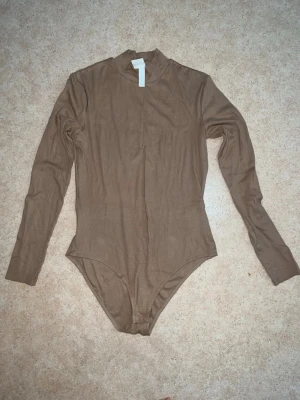 Beige långärmad body med hög hals - Säljer en beige långärmad body från H&M med hög hals. Bodyn är i mjukt, stretchigt material och har en enkel, stilren design utan synliga detaljer. Perfekt att styla med jeans eller kjol för en clean look.