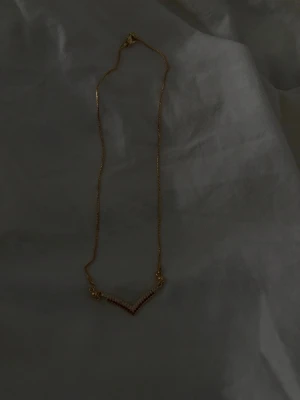 Guldfärgat halsband med pärldetaljer - Snyggt halsband i guldfärg med en tunn kedja och en v-formad hängare dekorerad med små pärlor. Perfekt accessoar för att lyfta din outfit med en elegant touch. Passar dig som gillar stilrena smycken med en twist.