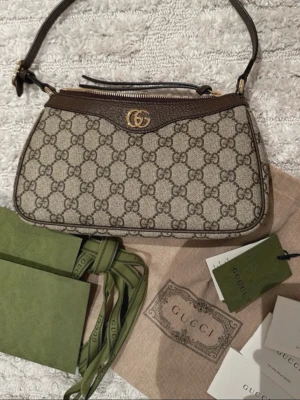 Gucci brun axelväska  - Säljer en snygg axelväska från Gucci med klassiskt GG-mönster i beige och brunt. Väskan har detaljer i brunt skinn, guldfärgad logga och dragkedja upptill. Perfekt storlek för det viktigaste och kommer med originalpåse och tags. 