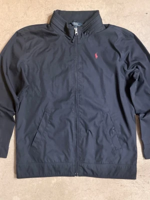 Polo Ralph Lauren windbreaker - Stilren svart jacka från Polo Ralph Lauren med dragkedja framtill och två snedställda fickor. Jackan har en röd broderad logga på bröstet och står ut med sin klassiska design. Perfekt för dig som gillar enkel men snygg streetstil.