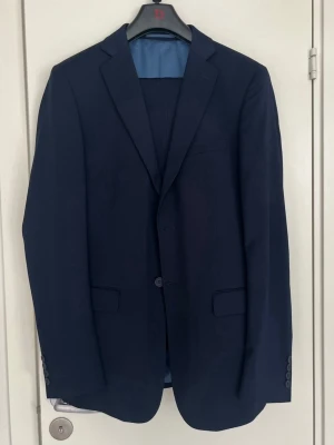 Mörkblå kostym med kavaj och byxor från dressmann - Snygg mörkblå kostym med klassisk kavaj och matchande byxor från dressmann. Kavajen har två knappar, smala slag och fickor med lock. Perfekt för dig som vill ha en stilren och tidlös look. Kostymen är helfärgad och har en modern passform.