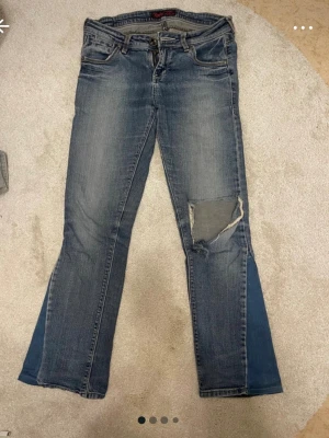 Blå bootcut jeans med patch - Säljer ett par blå bootcut jeans med slitningar och en stor patch på höger knä. Jeansen har klassisk femficksdesign och en snygg tvättad look. Perfekta för dig som gillar en avslappnad och trendig stil. (Dragkedjan sönder)