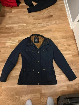 Marinblå quiltad jacka Hampton Republic - Snygg marinblå quiltad jacka från Hampton Republic med guldfärgade knappar och dragkedja. Jackan har lång ärm, flera fickor framtill och en brun innerfoder. Klassisk design med krage och detaljer i guld. Perfekt för dig som gillar stilren och tidlös look.