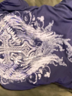 Blå croptop  med kors och vingar - Snygg blå croptop med stor vit tryck på ryggen som föreställer ett kors och änglavingar, dekorerad med glittriga detaljer.  Den är perfekt för dig som gillar statement-plagg med unika mönster.