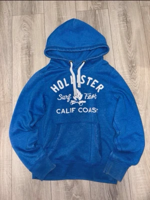 blå hoodie från Hollister - Blå, a hoodie från Hollister med vit text 'Surf Fest Calif Coast' och tryckt logga framtill. Klassisk kängur trainficka, vita dragsnören i huvan och ribbade muddar. Perfekt för chill dagar och streetwear vibes.