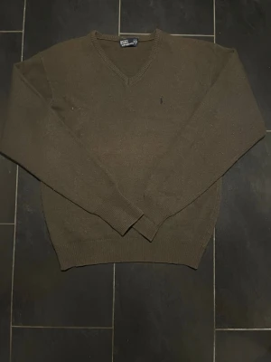 Olivgrön tröja från Polo Ralph Lauren - Olivgrön stickad tröja från Polo Ralph Lauren med V-ringning och diskret broderad logga på bröstet. Tröjan har långa ärmar och ribbade muddar vid ärmslut och nederkant. Perfekt för en stilren och avslappnad look.