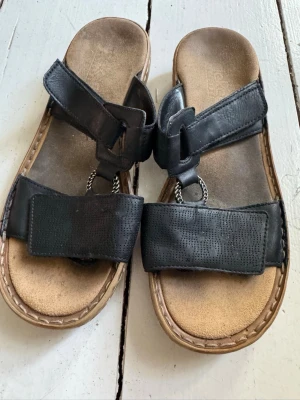 Sandaler från Rieker - Säljer ett par sandaler från rieker med breda remmar i svart läder och coola metallringar framtill. De är justerbara med kardborrband.  Sula i beige mockaimitation och kontrastsömmar runt kanten. Perfekta för chill dagar och enkel stil.