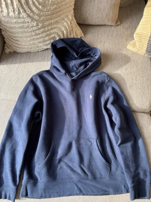Mörkblå hoodie från Polo Ralph Lauren - Säljer en mörkblå hoodie från Polo Ralph Lauren i storlek 18/20 Klassisk design med huva och stor magficka framtill. Materialet är mjukt och bekvämt, perfekt för en avslappnad look. Passar dig som gillar stilrena och tidlösa plagg.