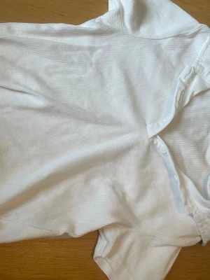 Vit ribbad t-shirt med krage - Säljer en vit ribbad t-shirt med korta ärmar och klassisk krage. Tröjan har knappar vid halsen för en clean och enkel look.