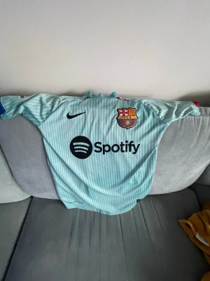 Barcelona bortatröja Nike ljusblå - Säljer en ljusblå FC Barcelona bortatröja från Nike med tunna vita ränder. Tröjan har klubbmärke och Nike-logga på bröstet samt Spotify som sponsortryck. Detaljer i rött och blått vid ärmslut och hals. Perfekt för dig som älskar fotboll och Barca.