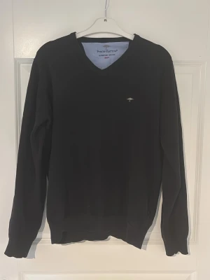 Svart tröja med V-ringning från Fynch-Hatton - Klassisk svart tröja från Fynch-Hatton med V-ringning och diskret broderad logga på bröstet. Tröjan har långa ärmar och ribbade muddar vid ärmslut och nederkant. Perfekt att bära över en skjorta för en stilren look.