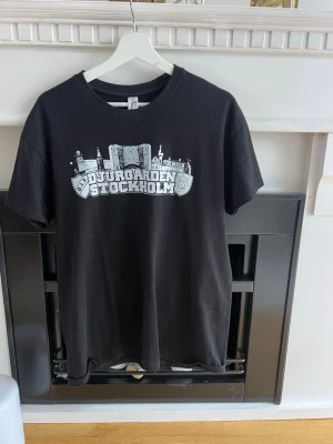 Svart Djurgården Stockholm t-shirt - Säljer en svart t-shirt med tryck av Djurgården Stockholm och stadssilhuett i vitt på bröstet. Klassisk passform med rund hals och korta ärmar. Perfekt för dig som vill representera Stockholm och Djurgården på ett stilrent sätt.