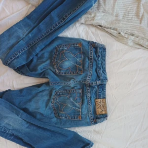 Blå jeans med broderade bakfickor - Säljer ett par blå jeans med unika broderade mönster på bakfickorna och kontrastsömmar. Jeansen har klassisk femficksdesign och ett snyggt läderpatch i midjan. Perfekta för dig som vill ha något extra till din denimlook. Innerbensläng: 77 Midjemått: 34 