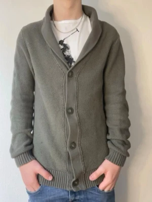Olivgrön cardigan från Lindbergh - Snygg olivgrön cardigan från Lindbergh i ekologisk bomull. Jackan har stora knappar framtill och en bred sjalkrage. Perfekt för lager på lager och ger en avslappnad men stilren look.