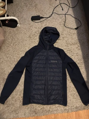 Mörkblå cardigan Peak Performance - Snygg mörkblå cardigan från Peak Performance med quiltad front och huva. Jackan har dragkedja framtill, två sidofickor och är tillverkad i en mix av slätt och vadderat material. Perfekt för blåsiga dagar och sportig stil.