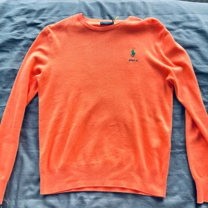 Orange tröja från Polo Ralph Lauren - Snygg orange tröja från Polo Ralph Lauren med klassisk rund halsringning och broderad grön logga på bröstet. Tröjan är långärmad och tillverkad i mjuk bomull, perfekt för alla årstider och nu till vintern ❄️. 