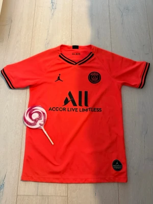 Röd PSG matchtröja Jordan - Snygg röd PSG matchtröja i samarbete med Jordan. Tröjan har svarta detaljer på krage och ärmslut, samt PSG-logga och Jordan-logga på bröstet. Tillverkad i lätt och ventilerande material, perfekt för fotboll eller träning.