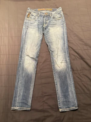 Jack & Jones jeans med slitningar - Säljer ett par ljusblå jeans från Jack & Jones med raka ben och snyggt slitna detaljer framtill och baktill. Klassisk femficksmodell med gylf och knapp. Perfekta för en avslappnad och trendig look.