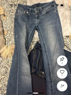 Blå bootcut jeans från True Religion - Säljer ett par klassiska blå bootcut jeans från True Religion med ikonisk vit kontrastsöm på bakfickan. Jeansen har låg midja, fem fickor och slitna detaljer framtill. Perfekta för en avslappnad och trendig look. Har lappen och kvitto kvar