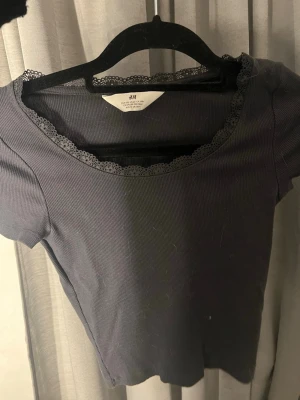 Mörkblå ribbad topp från H&M - Mörkblå ribbad topp från H&M med korta ärmar och söt spetskant runt halsringningen. Perfekt att styla med jeans eller kjol för en enkel men snygg look.