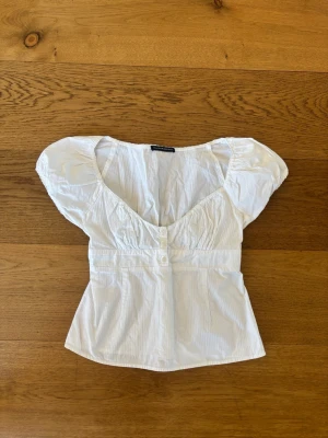 Vit blus från Brandy Melville - Supersöt vit blus från Brandy Melville med puffiga korta ärmar och v-ringad hals. Toppen har knappar framtill och markerad midja som ger en feminin siluett. Perfekt till jeans eller kjol för en fräsch look.