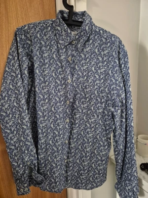 Blå paisleymönstrad skjorta Dressmann - Snygg blå skjorta från Dressmann med vitt paisleymönster. Tillverkad i en linnemix som känns lätt och fräsch. Skjortan har klassisk krage, knappar framtill och långa ärmar. Perfekt för dig som vill sticka ut lite extra.