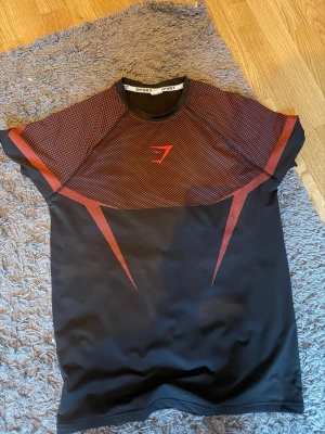 Svart och röd träningströja Gymshark - Snygg svart träningströja från Gymshark med röda grafiska detaljer och meshpartier över axlar och rygg. Tillverkad i ett stretchigt och snabbtorkande material som passar perfekt till gymmet eller löpning. Tight passform och korta ärmar.