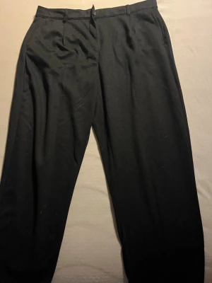 Svarta kostymbyxor med raka ben - Snygga svarta kostymbyxor med klassisk rak passform och hällor för skärp. Byxorna stängs med dragkedja och knapp. Perfekta för en clean och stilren look. Materialet känns mjukt och följsamt.