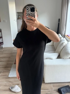 Svart enkel långklänning - Svart långklänning med rund halsringning och korta ärmar. Klänningen har en rak passform och är tillverkad i ett mjukt material som ger en avslappnad look. Perfekt för dig som gillar minimalistisk stil.