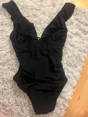 Svart baddräkt med volangaxel - Stilren svart baddräkt med djup v-ringning och breda axelband med volangdetaljer. Klassisk modell som ger en snygg siluett och passar perfekt till stranden eller poolen. Tillverkad i stretchigt material för skön passform.