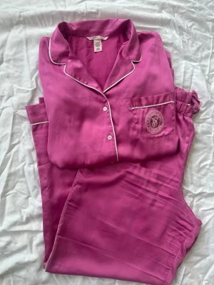 Rosa satinpyjamas från Victoria's Secret - Snygg rosa pyjamas i glansigt satinmaterial från Victoria's Secret. Setet har vita kontrastsömmar, knappstängning och en bröstficka med VS-logga. Perfekt för mysiga kvällar hemma och ger en lyxig känsla.