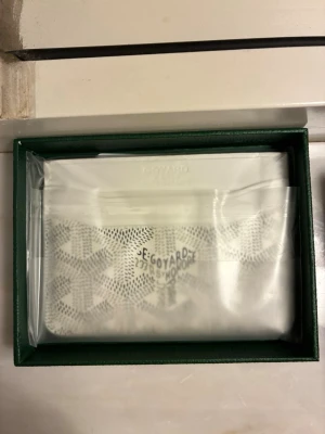 Vit plånbok i box - Snygg vit plånbok med det klassiska mönstret och loggan tryckt på framsidan. Plånboken är tillverkad i canvas med detaljer i läder och levereras i grön box med silverfärgad logga. Perfekt för dig som gillar exklusiva accessoarer.