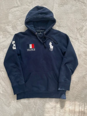 Ralph Lauren - Snygg marinblå hoodie från Ralph Lauren med broderad fransk flagga och texten FRANCE på bröstet. Stor vit Polo-logga på ena sidan och siffran 3 på ärmen. Klassisk känguruficka och dragsko i huvan. Perfekt för dig som gillar sportig och stilren look.