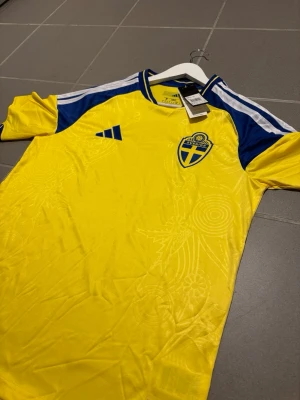 Svensk landslagströja  - Säljer en svensk landslagströja från Adidas i gult med blå detaljer och vita ränder på axlarna. Tröjan har broderad svensk landslagssköld på bröstet och Adidas-logga. Tillverkad i lätt och ventilerande material, perfekt för fotboll eller träning.