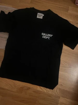 Svart t-shirt från Gallery Dept. - Snygg svart t-shirt från Gallery Dept. med vit logga tryckt på bröstet. Klassisk passform och rund halsringning. Perfekt till jeans eller shorts för en clean och enkel stil.