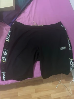 Svarta EA7 shorts med logga - Svarta shorts från EA7 Emporio Armani med tydliga loggor längs sidorna och framtill. Modellen har elastisk midja med snörning och är tillverkad i mjukt material, perfekt för sommaren och för dig som vill ha en avslappnad och sportig stil. Användt några gånger liten fläck som ni ser på bild 2 😊hör av er vid frågor 