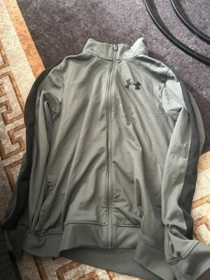 Grå ziptröja från Under Armour - Snygg grå ziptröja från Under Armour med hög krage och hel dragkedja framtill. Tröjan har två sidofickor och är tillverkad i ett mjukt, stretchigt material som passar perfekt till träning eller chill. Diskret logga på bröstet för en sportig look.