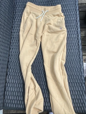 Beige mjukisbyxor från GANT - Snygga beige mjukisbyxor från GANT med dragsko i midjan och ribbade muddar vid bensluten. Byxorna har fickor på sidorna och en liten GANT-logga broderad på ena benet. Tillverkade i mjukt bomullsmaterial som känns riktigt skönt mot huden.