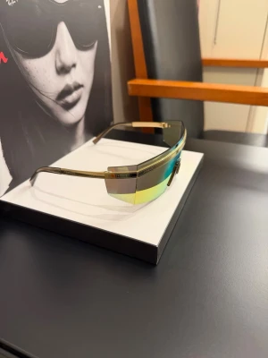 Versace futuristiska solglasögon - Säljer ett par futuristiska Versace solglasögon med spegelglas i guld och gröna toner. Modellen har en skarp, rektangulär form med bred front och tunna metallbågar. Perfekta för dig som vill sticka ut med en modern och edgy look.