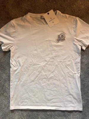 Vit Moncler t-shirt med logga - Snygg vit t-shirt från Moncler med rund hals och två broderade loggor på bröstet. Tillverkad i mjuk bomull och har en klassisk passform med korta ärmar. Perfekt för en clean och stilren look.