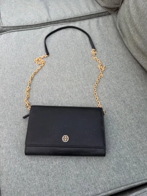Svart axelväska från Tory Burch - Snygg svart axelväska från Tory Burch i slätt läder med guldfärgad kedja och logga framtill. Väskan har flera fack, inklusive dragkedjefack och kortplatser, samt blått innerfoder. Perfekt för dig som vill ha en stilren och praktisk väska.
