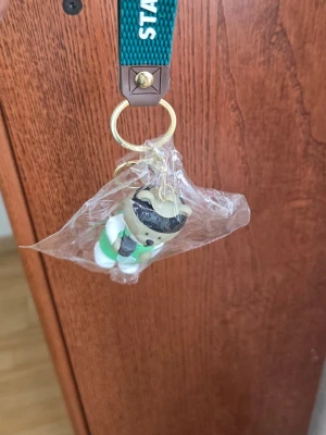 Starbucks nalle nyckelring - Söt nyckelring med en liten nalle i Starbucks-uniform. Nallen har grön förkläde med Starbucks-logga, vit tröja och svart keps. Perfekt accessoar till väskan eller nyckelknippan för dig som älskar Starbucks och gulliga detaljer.