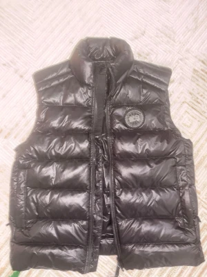  Canada Goose crofton väst - Snygg svart dunväst från Canada Goose med hög krage och klassisk logotyp på bröstet. Västen har en quiltad design och dragkedja framtill. Perfekt för lager-på-lager och håller dig varm under kyliga dagar.