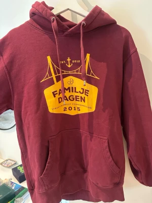 Vinröd hoodie - Vinröd hoodie med tryck i gult framtill från Familjedagen 2015. Klassisk känguruficka, dragsko i huvan och ribbade muddar. Skön och avslappnad passform, 
