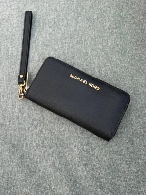 Svart plånbok Michael Kors - Stilren svart plånbok från Michael Kors i läder med guldfärgade detaljer och dragkedja runtom. Plånboken har flera fack för kort, myntfack med dragkedja och avtagbar handledsrem. Perfekt för dig som vill ha både stil och funktion.